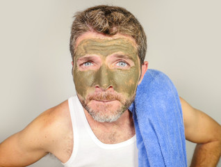 young messy funny man looking at himself horrified in bathroom mirror with green cream on his face applying beauty facial mask product