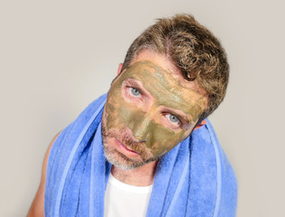 young messy funny man looking at himself horrified in bathroom mirror with green cream on his face applying beauty facial mask product