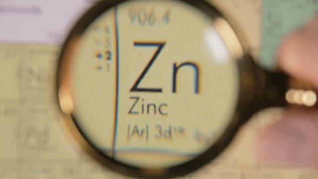 A hand moving a lens magnifier over a defocused board (a periodic table) and revealing the chemical element name, symbol and scientific properties of Zn, zinc.

