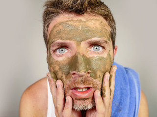 young messy funny man looking at himself horrified in bathroom mirror with green cream on his face applying beauty facial mask product