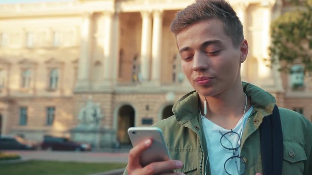 Close up face handsome young man use phone in headphone look around standing on steet city smile cell sunlight happy gadget technology hipster slow motion portrait