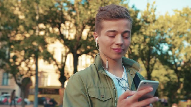 face handsome young man use phone in headphone look around standing on steet city smile cell sunlight happy gadget technology hipster slow motion portrait close up