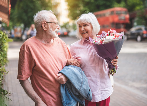 Side view of happy couple standing on street. Elderly female is holding beautiful bunch of flowers presented by male. They are looking at each other and smiling
