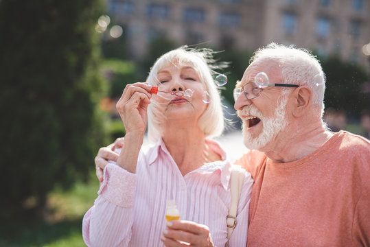 Cheerful lady holding bottle with soap water and blowing bubbles on street. Exited mature male is hugging and looking at her with content. They are embracing each other with pleasure