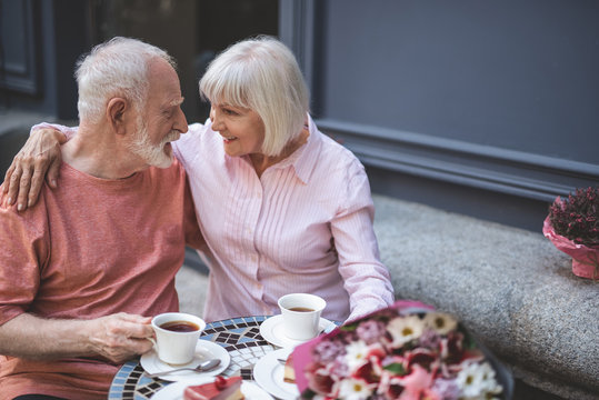 Side view of delighted man and woman drinking tea outside on date. They are looking at each other with love while elderly female is embracing her spouse