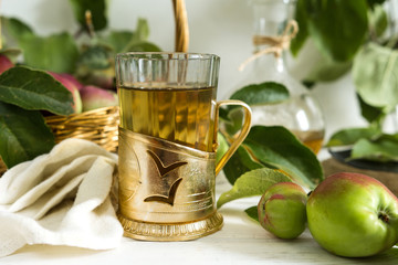 Apple tea in a glass goblet and a decanter and a basket with ripe apples on a wooden table in...