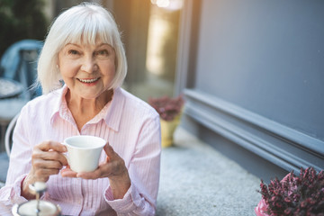 Waist up portrait of happy elderly woman sitting at table in cafe and holding cup of tea. She is...