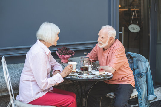 Side view of attentive man sitting at table with old woman and listening to her. Elderly lady is talking with joy while drinking tea and eating cake. They are enjoying evening outdoors together