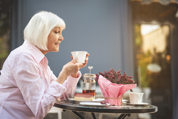 Side view profile of happy senior lady holding cup of hot drink and smiling. She is enjoying evening dinner surrounded by city nature