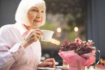 Side view of joyful senior woman sitting at table and holding cup of tea in hand.  She is enjoying evening outdoors with content