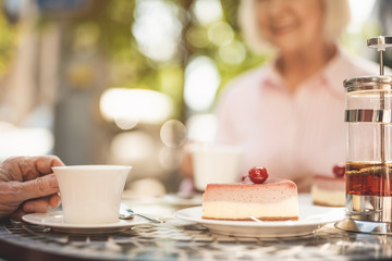 Focus close up of mature male hand holding cup of hot drink. Piece of delicious cake is lying beside. Old lady is on background