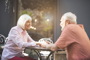 Side view of smiling senior lady and man sitting at table outside. They are looking at each other with joy and holding hands with delight