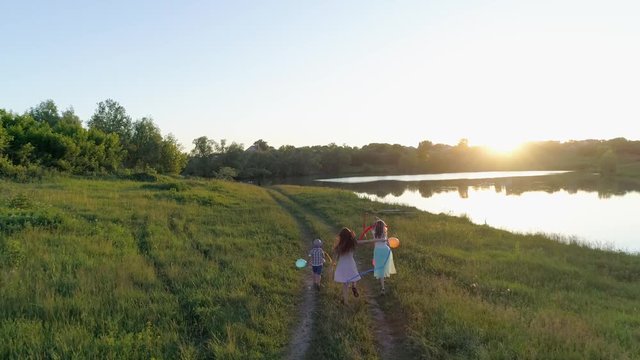 drone view, cheerful children with colored ribbons and balls in hands are running on green meadow near river