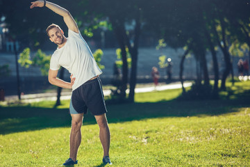 Full length portrait of smiling attractive man doing exercises outdoors. He is bending with raised...