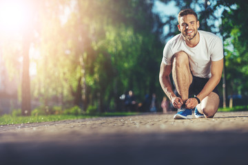 Full length portrait of smiling athlete kneeling and fixing shoelaces outdoors. He is joyfully looking at camera while listening to music with earphones. Copy space in left side