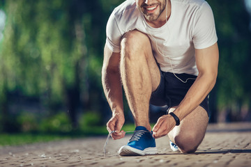 Full length of smiling athlete kneeling on ground and tying snickers with joy. He is happy and delighted to workout outdoors. Copy space in left side