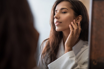 I am so beautiful. Portrait of pleased pretty woman touching face with hand while looking at mirror