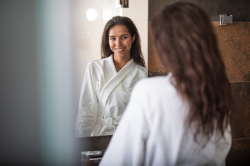 Portrait of beaming pretty girl looking at mirror. She wearing cozy bathrobe while locating in apartment