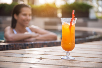 Close up glass of appetizing orange cocktail locating opposite outgoing female. She relaxing in water during hot day