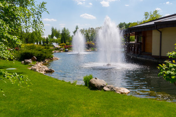 Refreshing transparent fountains in the middle of a pond against a blue sky with a green lawn on one side and a restaurant on the other.