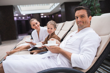 Waist up portrait of delighted man and woman sitting on deckchairs with kid. They are holding cups with hot drink to get warm after hydrotherapy in spa