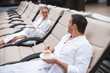 Side view of smiling male and female sitting on deckchairs and drinking tea. Young people are communicating while relaxing with joy and delight