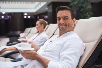 Focus on waist up portrait of joyful male sitting on deckchair with cup of favorite hot drink. He is enjoying relaxation time. Young woman in bathrobe is on background
