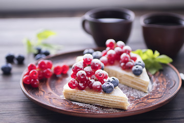 Two pieces of puff cake with fresh berries and sugar powder on brown plate and two brown cups of coffee on dark wooden table.