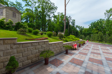 A house with a green lawn located on a slope. Under it, from the tiles laid alley with benches, on which women sit