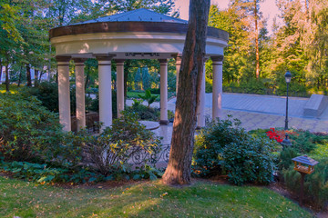 Decorative gazebo with white columns and forged fence against the background of green plants