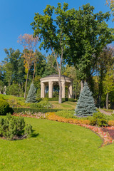 A massive stone pavilion with wide columns and a black roof against a green lawn and tall trees