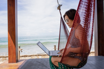 Asian business woman sit on hammock and works on laptop at beachfront during vacation