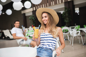 Focus on waist up portrait of happy woman sitting at table in cafe. She is holding glass with cocktail and looking sideways with delight. Young man with drink on background