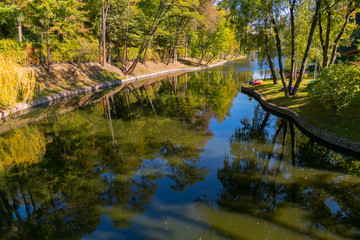 People who are resting on a clear summer day on the shore of a pond with a beach lined with stone and a beautiful view of the water with reflecting trees in it.