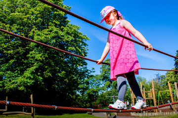 Young girl crossing a rope bridge in a playground with a blue sky