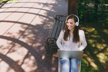 Young successful businesswoman in light casual clothes. Woman working on modern laptop pc computer, listen music in headphones on head in street outdoors. Mobile Office. Freelance business concept.