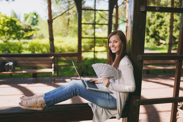 Obraz premium Young successful businesswoman holding bundle of dollars, cash money. Woman working on modern laptop pc computer in city park in street outdoors on nature. Mobile Office. Freelance business concept.