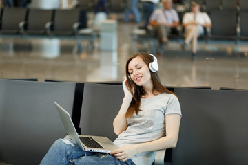 Young relaxed traveler tourist woman with headphones listening music working on laptop, wait in lobby hall at international airport. Passenger traveling abroad on weekends getaway. Air flight concept.