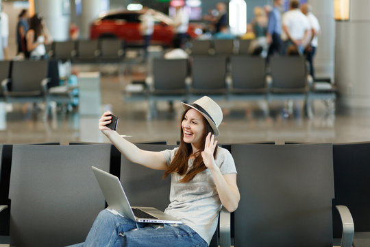 Young smiling traveler tourist woman working on laptop, doing selfie on mobile phone waiting in lobby hall at international airport. Passenger traveling abroad on weekends getaway. Air flight concept.