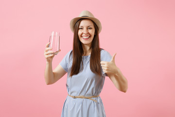 Portrait of young beautiful woman in blue dress, hat holding and drinking clear fresh pure water from glass isolated on pink background. Healthy lifestyle, people, sincere emotions concept. Copy space