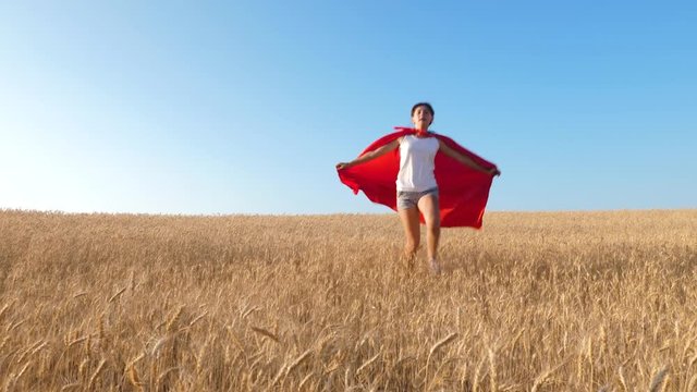 Superhero girl running across field with wheat against blue sky and smiling.