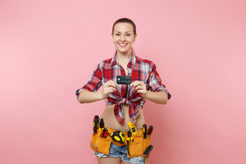 Young smiling handyman woman in plaid shirt, denim shorts, kit tools belt full of variety instruments holding bank credit card isolated on pink background. Female doing male work. Renovation concept.