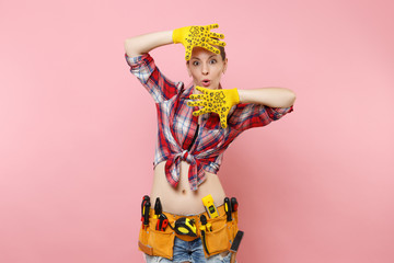 Strong young fun handyman woman in plaid shirt, denim shorts, yellow gloves, kit tools belt full of variety useful instruments isolated on pink background. Female doing male work. Renovation concept.