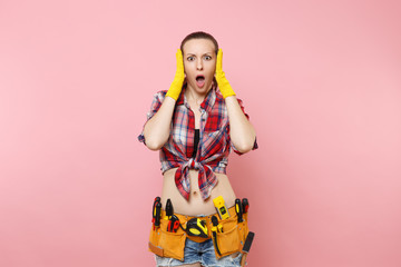 Shocked stress handyman woman in plaid shirt, denim shorts, yellow gloves, kit tools belt full of variety useful instruments isolated on pink background. Female doing male work. Renovation concept.