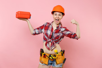 Strong young woman in orange helmet, plaid shirt, denim shorts, kit tools belt full of instruments, toolbox showing biceps, muscles isolated on pink background. Female in male work. Renovation concept