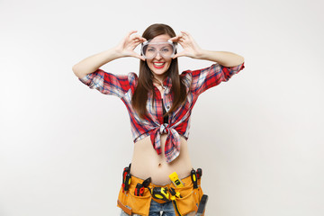 Strong young handyman woman in plaid top shirt, denim shorts, kit tools belt full of instruments in protective goggles isolated on white background. Female in male work. Copy space for advertisement.