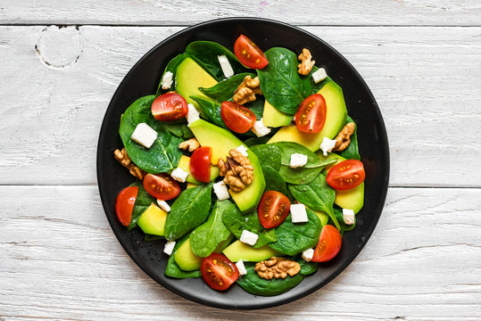 avocado and spinach salad with tomatoes cherry, feta cheese and walnuts in a plate on rustic wooden table. top view