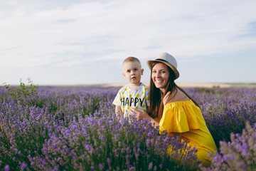 Young woman in yellow dress walk on purple lavender flower meadow field background, rest, have fun, play with little cute child baby boy. Mother, small kid son. Family day, parents, children concept.