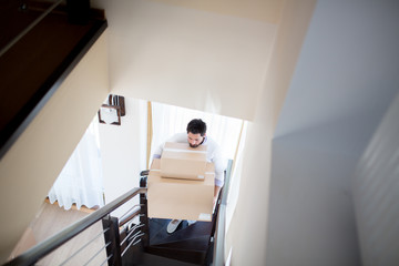 Young man with big and smaller boxes going upstairs inside new house after relocation