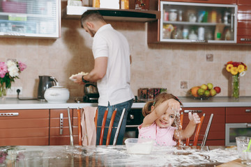 Little kid girl plays with flour while daddy cooking in kitchen at table. Happy family dad, child daughter cooking food in weekend morning at home. Father's day holiday. Parenthood, childhood concept.